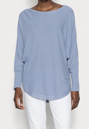 Kvinna iklädd en löst sittande ljusblå stickad tröja med sänkt axel och vita byxor mot en enkel ljus bakgrund.