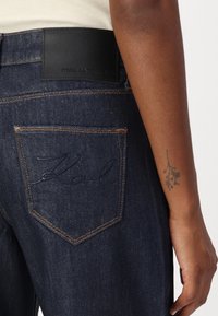Dunkelblaue Denim-Jeans mit einem schwarzen Lederlogo-Patch am Bund. Signatur "Karl" in blauer Stickerei auf der Gesäßtasche.
