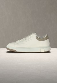 Άσπρο παπούτσι τύπου sneaker χαμηλού γυρισμού με μπεζ ταμπελάκι στη φτέρνα, κλείσιμο με κορδόνια, ανάγλυφο λογότυπο στο πλάι και παχύ λευκό πέλμα με μπεζ λεπτομέρειες στην εξωτερική σόλα.