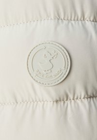 Jachetă puf, bej deschis, cu textură matlasată. Prezintă un logo circular, alb-crem "Save the Duck" cu un grafic de rață în centru.