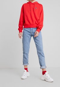 Persona vistiendo una sudadera roja con capucha, jeans claros arremangados, calcetines rojos y zapatillas blancas gruesas, de pie contra un fondo gris plano.