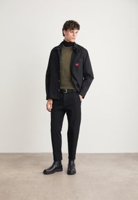 Black shirt jacket with a red logo patch over a green turtleneck, paired with black tapered trousers and black ankle boots.