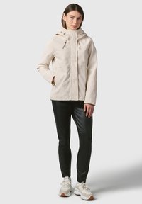 Lichtbeige hooded jack met voorrits, twee zakken en een trekkoord. Draagt met zwarte slim-fit broek en beige sneakers.