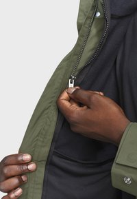 Olivengrøn jakke med en glat overflade, lynlåslukning og et blødt sort fleecefore, der er synligt. En hånd griber fat i lynlåsens træk.