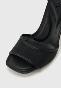 Μαύρο σανδάλι ανοιχτής μύτης με φαρδιά ιμάντα από δίχτυ και επενδεδυμένο πέλμα, που διαθέτει ελικοειδή υποστήριξη καμάρας και σχεδιασμό angular στην φτέρνα.