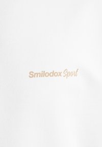 Hvid bomulds t-shirt med beige "Smilodox Sport" tekst. Glat tekstur med et simpelt, rent design, uden yderligere mønstre eller accenter.