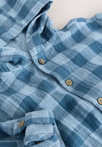 Chemise à manches longues à carreaux bleus et gris clair avec des boutons en bois, partiellement déboutonnée et froissée sur une surface plane.