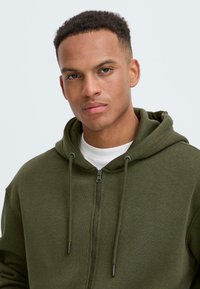 Jonge man met kort haar draagt een olijfgroene hoodie met rits over een wit T-shirt en kijkt recht in de camera met een neutrale uitdrukking.
