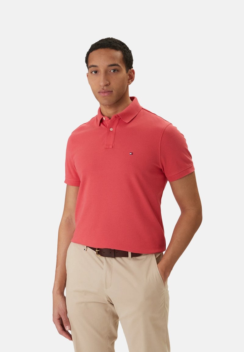 Homme portant un polo corail à manches courtes et un pantalon beige avec une ceinture marron foncé, debout avec une main dans la poche devant un fond uni.