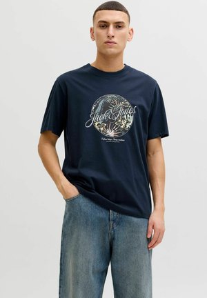 Young man wearing a navy blue Jack & Jones t-shirt with floral circular design and blue jeans, standing against a plain light background.