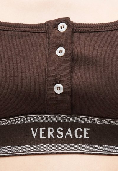 Καφέ τοπ από ριμπ ύφασμα με τρία λευκά κουμπιά και μαύρη ελαστική ταινία στο κάτω μέρος με την επιγραφή "VERSACE" σε λευκά γράμματα.