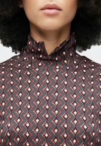 Tuva un kakls, valkājot melnu augstu apkakli ar sarkaniem, baltiem un pelēkiem dimanta ģeometriskiem rakstiem, ar cirtainiem tumšiem matiem, kas ietver seju.