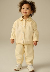 Veste à rayures jaunes et blanches avec boutons-pression et col rond, assortie à un pantalon et des baskets légères, portée par un enfant.