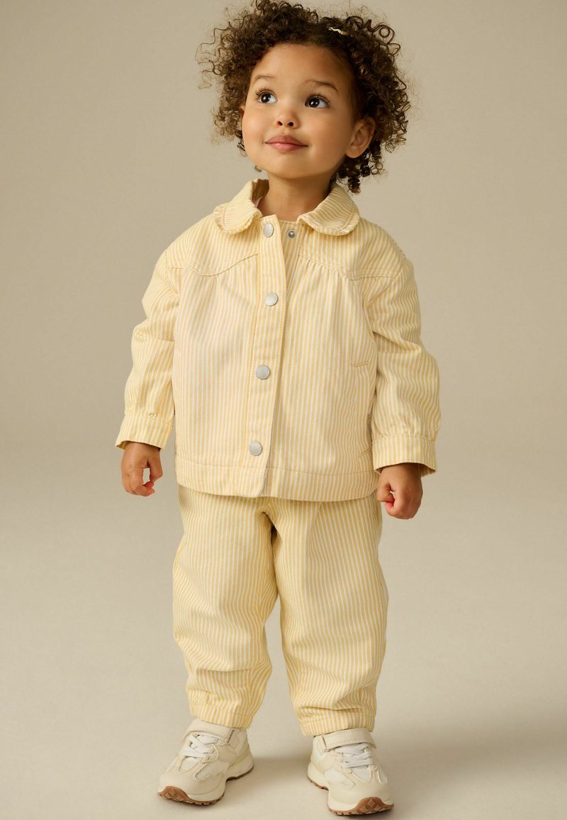 Veste à rayures jaunes et blanches avec boutons-pression et col rond, assortie à un pantalon et des baskets légères, portée par un enfant.