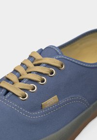 Μπλε αθλητικό παπούτσι Vans με μπεζ κορδόνια, λευκή ραφή, μεταλλικά πατζούρια και καφέ σόλα, εμφανίζεται σε κοντινό πλάνο.