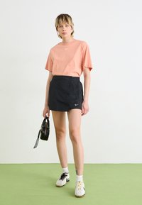 Peach cotton t-shirt with logo, black button-up mini skirt, white athletic shoes with yellow laces, and small black handbag. Green floor.