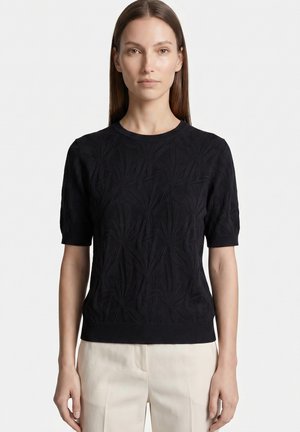 Frau mit glatten braunen Haaren, die ein schwarzes, strukturiertes Kurzarm-Top und hellbeige Hosen trägt und vor einem schlichten Hintergrund steht.