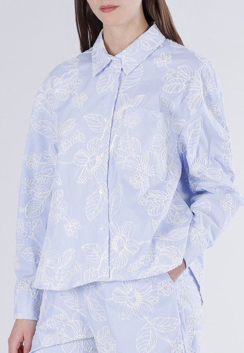 Vrouw draagt een lichtblauw gestreept overhemd en broek met witte bloemenborduursels, hand in de zak, staand tegen een effen achtergrond.