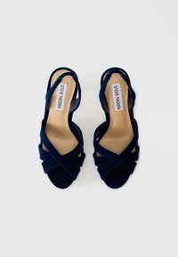 Tumši zila samta sandales ar atvērtu purngalu, krustveida siksnām, gludu bēšu iekšpusi un nedaudz izliekta papēža. Uz etiketes ir rakstīts "Steve Madden."