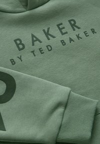Zeleni pamučni dukserica s rebrastim manžetama. Ima istaknut otisak: "BAKER BY TED BAKER" u tamnozelenoj boji na prednjoj strani. Glatka tekstura.