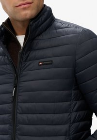 Navy puffer jacket with horizontal quilted texture, high collar, and zip closure. Features a small black and red brand logo on the chest.