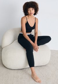 Top corto a coste nero abbinato a joggers neri larghi. Il modello è seduto su una sedia rotonda di colore chiaro e texture, su un morbido tappeto beige.