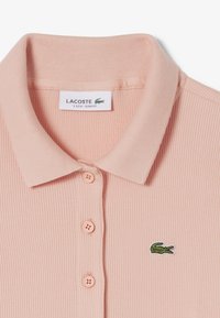 Polo in tessuto a costine rosa chiaro con colletto, bottoni solidi e logo coccodrillo Lacoste verde ricamato sul petto.