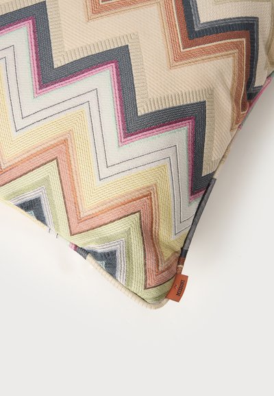 Coussin carré avec motif zigzag multicolore en beige, orange, vert, rose, noir et gris, avec une petite étiquette en tissu orange.