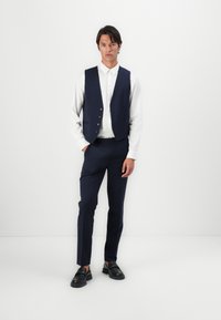 Marineblauer Weste mit fünf Knöpfen über einem weißen Hemd, kombiniert mit passenden marineblauen Hosen und schwarzen Schuhen, im schlanken Schnitt.
