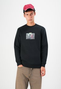 Black sweatshirt with a crew neck and ribbed cuffs, featuring a graphic print of a brick wall and a red telephone booth. Worn with beige pants.