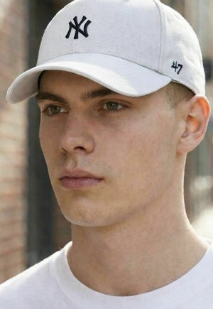 Junger Mann trägt eine weiße Baseballkappe mit schwarzem "NY"-Logo und der auf der Seite gestickten Zahl "47", blickt mit neutralem Ausdruck nach vorn.