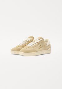 Beige suède sneakers met witte veters en accenten, voorzien van een getextureerde zool en een merkdetail aan de zijkant. Klassiek laag ontwerp.