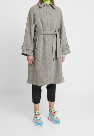 Manteau trench en chevrons gris avec une taille ceinturée, un large col et des accents en boutons, associé à des sneakers colorées et un pantalon noir cropped.
