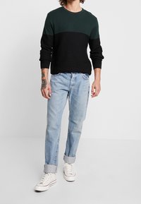Pull côtelé noir et vert, col rond, associé à un jean bleu clair avec revers et des baskets blanches. Les deux vêtements ont une coupe décontractée.