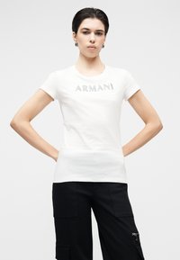 T-shirt branca de algodão com mangas curtas e logótipo "ARMANI" prateado. O corte é justo e o decote é redondo.