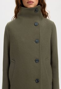 Olivenfarbener Wollmantel mit hohem Kragen und fünf schwarzen Knöpfen. Verfügt über seitliche Taschen und einen weichen, strukturierten Stoff.