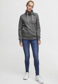 Felpa grigia con colletto a imbuto, stringhe regolabili e accessori in legno. Abbinata a jeans skinny blu e sneakers bianche.