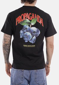 Uomo che indossa una maglietta nera con la scritta "PROPAGANDA" sopra mirtilli fatti di teschi e foglie verdi, e "FINEST SELECTION" sotto.