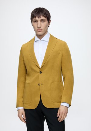 Mand iført sennepsgul blazer over hvid skjorte og sorte bukser, stående mod en ensfarvet hvid baggrund.