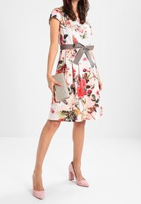 Floral dress with short sleeves, light pink fabric, cinched at the waist with a grey ribbon. Paired with a silver clutch and pink heels.