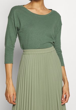 Kobieta w jasnozielonym topie z długim rękawem i szerokim dekoltem, zestawionym z wysoką talią, plisowaną, bladozieloną spódnicą.