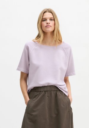 Kobieta o długich blond włosach, ubrana w jasnofioletową bluzkę z krótkim rękawem i wysokim stanem brązowych spodni, z rękami w kieszeniach.