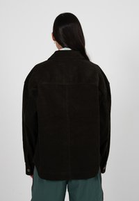 Frau mit langem dunklem Haar, die eine dunkelbraune Cordjacke und türkise Hose trägt, von hinten vor einem einfarbigen Hintergrund zu sehen.