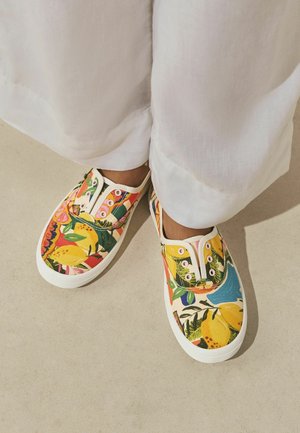 Kojos dėvint baltas kelnes ir slip-on stiliaus sportbačius su ryškiais citrusinių vaisių ir lapų raštais ant šviesios grindų spalvos.