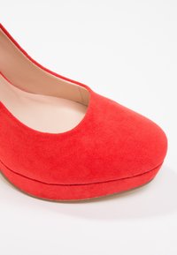 Chaussure à talon haut en daim rouge avec un bout arrondi et une doublure intérieure beige sur fond blanc.