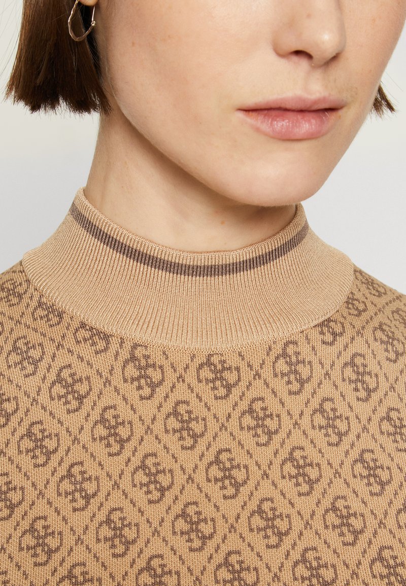Maglione beige lavorato a maglia con colletto alto a costine e motivo geometrico a logo in marrone. La vista ravvicinata mette in evidenza texture e dettagli del design.