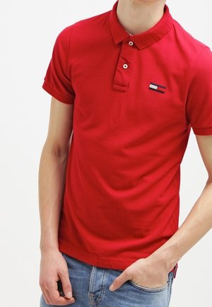 Man wearing a red short-sleeve polo shirt with a small logo on the chest, paired with blue jeans, one hand in pocket.