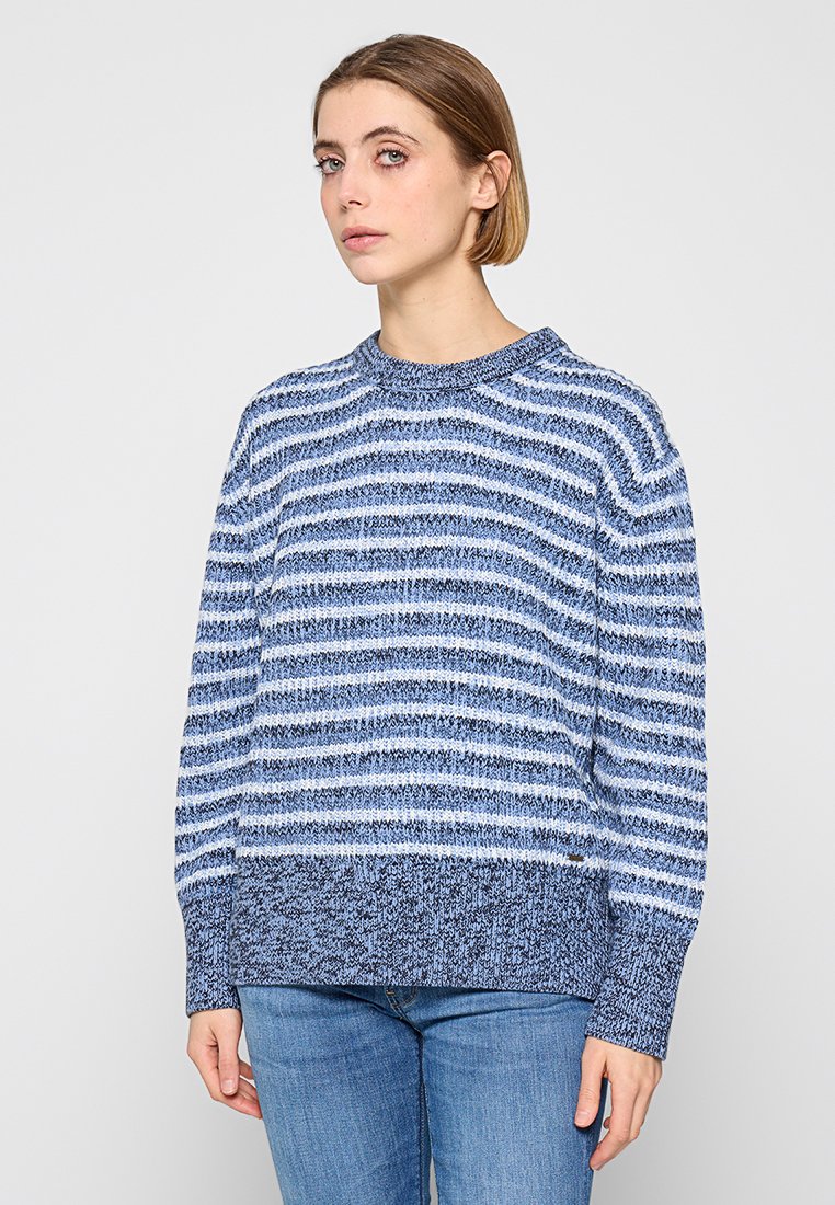 Suéter de punto a rayas azules con escote redondo, que presenta diferentes tonalidades de azul y un dobladillo azul texturizado. Combinado con jeans de corte claro.