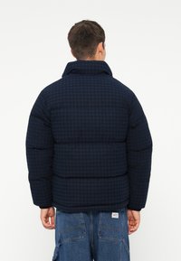 Marineblå pufferjakke med rutete mønster, høy krage og elastiske mansjetter. Stylet med løstsittende denimbukser.