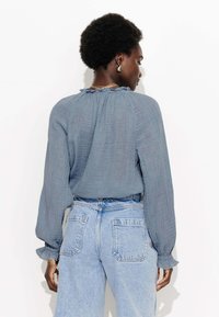 Blouse longue à manches longues bleu clair texturé avec un décolleté froncé et un col à volants. Portée avec un jean en denim léger taille haute.